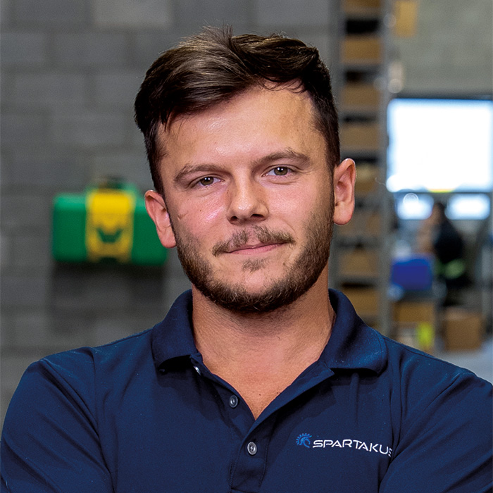 Professional headshot of a man in a blue Spartakus polo shirt, industrial background.